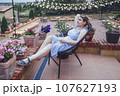 Young woman with glass in her hand is resting on terrace of house. Attractive blonde girl is sitting in wicker chair against background of evening cafe with garlands of light bulbs and pots of flowers 107627193