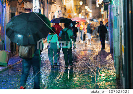 People walk along a narrow European street with umbrellas in the rain - evening and different colored lights of the night city People walk along a narrow European street with umbrellas in the rain - evening and different colored lights of the night city 107628525