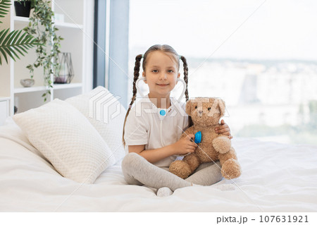Pretty kid with toy stethoscope examining teddy bear in bed 107631921
