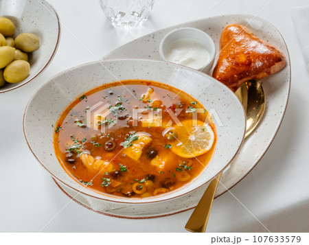 Solyanka soup with meat and olives garnished with a slice of lemon. Traditional Russian meat soup Solyanka in bowl on restaurant table 107633579