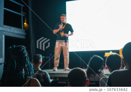 Male speaker standing on stage near big screen and giving presentation to participants sitting in dark hall during business seminar 107633682