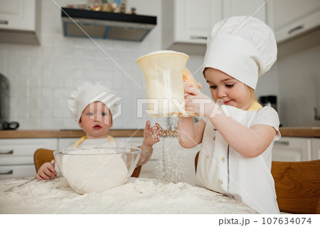 Adorable girl in chefs hat and apron sift flour with a sieve at the kitchen 107634074