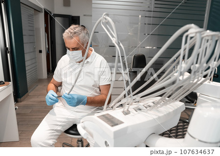 A dentist sits behind a dental chair and prepares tools for dental treatment. A senior, experienced male doctor works in a dental treatment and prosthetics center. 107634637