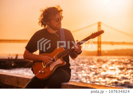 Hipster street musician in black playing electric guitar in the street on sunset on embankment with 25th of April bridge in background. Lisbon, Portugal Hipster street musician in black playing electric guitar in the street on sunset on embankment with 25th of April bridge in background. Lisbon, Portugal 107634824
