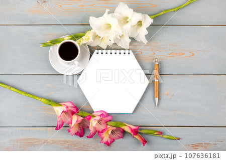 Composition with opened blank notepad and pen, cup of black coffee, white and pink gladiolus flower on blue wooden table. Festive office desktop concept. Morning coffee cup. Composition with opened blank notepad and pen, cup of black coffee, white and pink gladiolus flower on blue wooden table. Festive office desktop concept. Morning coffee cup. 107636181