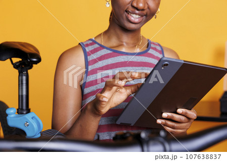 Active African American female cyclist browsing for bicycle repair solutions on her digital tablet. Sporty black woman using smart device to research bike adjustments for expert maintenance. Active African American female cyclist browsing for bicycle repair solutions on her digital tablet. Sporty black woman using smart device to research bike adjustments for expert maintenance. 107638837
