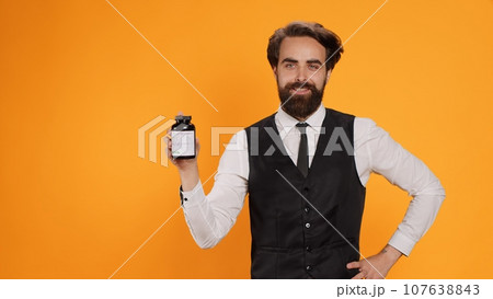 Fine dining butler holds vitamins jar on camera, presenting bottle of medical supplements while he works as waiter. Professional worker recommending medicaments pills in studio, catering industry. 107638843