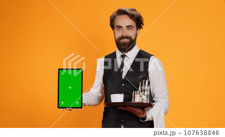 Friendly waiter shows tablet with greenscreen on camera, presenting blank copyspace mockup template. Experienced butler in suit holding device with isolated empty display and food tray. 107638846