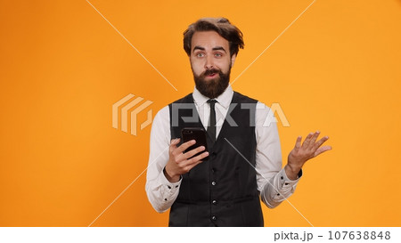 Restaurant employee messaging on duty and using phone at work, serving clients with meal orders. Classy waiter standing against yellow background in studio, checking smartphone social media apps. 107638848