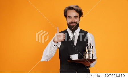 Accomplished valet gives thumbs up in studio, displays agreement sign and is enthusiastic on camera. Before serving cup of coffee, young employee with waiter profession indicates approbation. 107638850