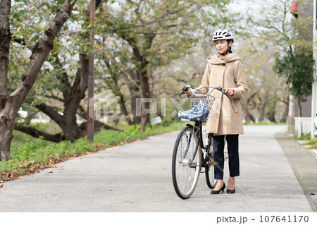 川沿いをリュックサックを背負ってキャメルロングコートを着て自転車を引いて歩く若い女性 川沿いをリュックサックを背負ってキャメルロングコートを着て自転車を引いて歩く若い女性 107641170