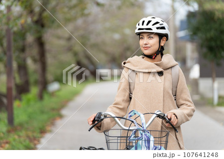 川沿いをリュックサックを背負ってキャメルロングコートを着て自転車を引いて歩く若い女性 上半身 107641248