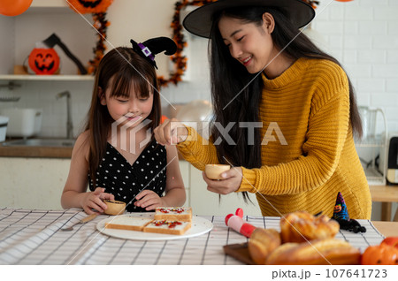 A cute young Asian girl is helping her mom prepare Halloween desserts in the kitchen. A cute young Asian girl is helping her mom prepare Halloween desserts in the kitchen. 107641723