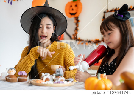 A beautiful Asian mom enjoys making Halloween cupcakes with her young daughter in the kitchen. 107641743