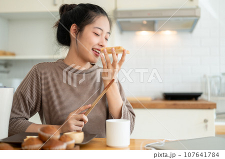A beautiful and happy young Asian woman is enjoying her toast in the kitchen, having breakfast A beautiful and happy young Asian woman is enjoying her toast in the kitchen, having breakfast 107641784