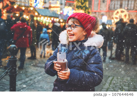 Little cute preschool girl drinking hot children punch or chocolate on German Christmas market. Happy child on traditional family market in Germany, Laughing kid in colorful winter clothes Little cute preschool girl drinking hot children punch or chocolate on German Christmas market. Happy child on traditional family market in Germany, Laughing kid in colorful winter clothes 107644646