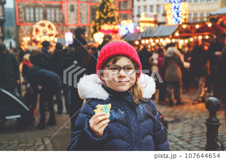 Little cute preschool girl eating gingerbread cookie on German Christmas market. Happy child on traditional family market in Germany, Laughing kid in colorful winter clothes Little cute preschool girl eating gingerbread cookie on German Christmas market. Happy child on traditional family market in Germany, Laughing kid in colorful winter clothes 107644654