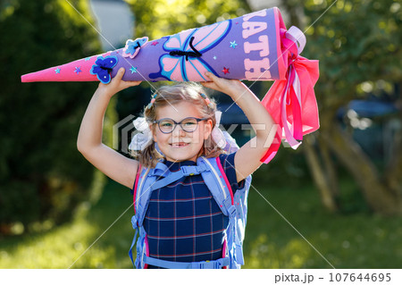 Happy little kid girl with eye glasses with backpack or satchel and big school bag or gift cone traditional in Germany for the first day of school. Healthy adorable child outdoors. Back to school 107644695