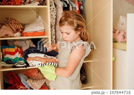 Little school girl standing by wardrobe with clothes. Preschool child putting shirts away and making decision for shirt to wear. Children get dressed in the morning for school. Little school girl standing by wardrobe with clothes. Preschool child putting shirts away and making decision for shirt to wear. Children get dressed in the morning for school. 107644718