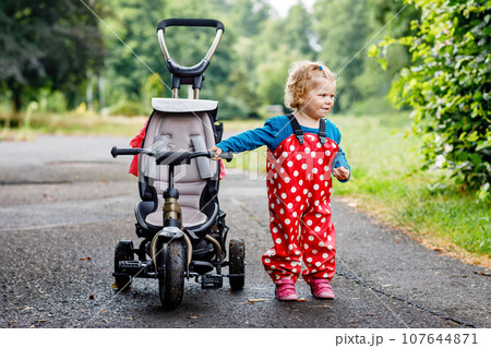 Cute adorable toddler girl sitting on pushing bicycle or tricycle. Little baby child going for a walk with parents cold cloudy day. Happy healthy kid in colorful clothes Cute adorable toddler girl sitting on pushing bicycle or tricycle. Little baby child going for a walk with parents cold cloudy day. Happy healthy kid in colorful clothes 107644871