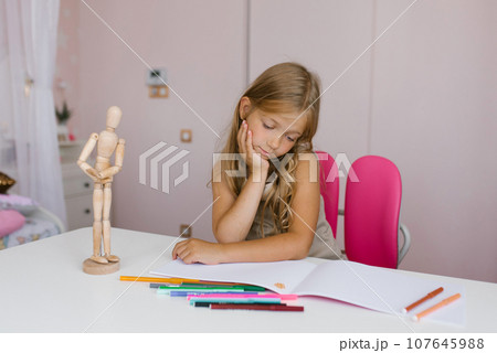 Seven year old schoolgirl is doing homework at her desk at home or at school, drawing with felt-tip pens, looking thoughtfully into a notebook Seven year old schoolgirl is doing homework at her desk at home or at school, drawing with felt-tip pens, looking thoughtfully into a notebook 107645988