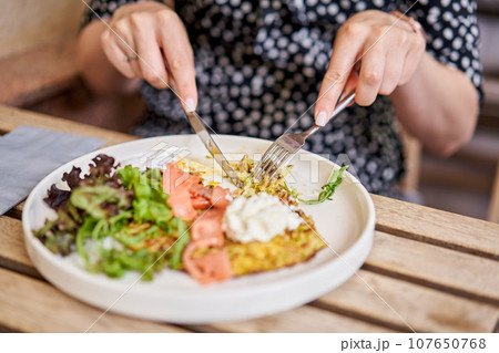 Woman cuts Zucchini pancakes, with avocado, cheese cream, salmon and egg . Healthy breakfast, protein. Restaurant dish. Breakfast in cafe on a sunny morning, on the summer veranda 107650768