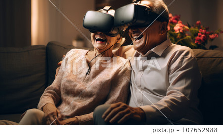 Senior mature couple wearing virtual reality headsets sits on the sofa having fun together. Happy retired people using modern VR goggle glasses. New trends and technology concept for all ages. Senior mature couple wearing virtual reality headsets sits on the sofa having fun together. Happy retired people using modern VR goggle glasses. New trends and technology concept for all ages. 107652988