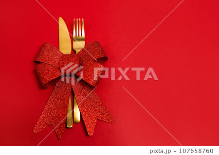Christmas table place setting. Golden fork and knife with red bow on red background, copy space Christmas table place setting. Golden fork and knife with red bow on red background, copy space 107658760