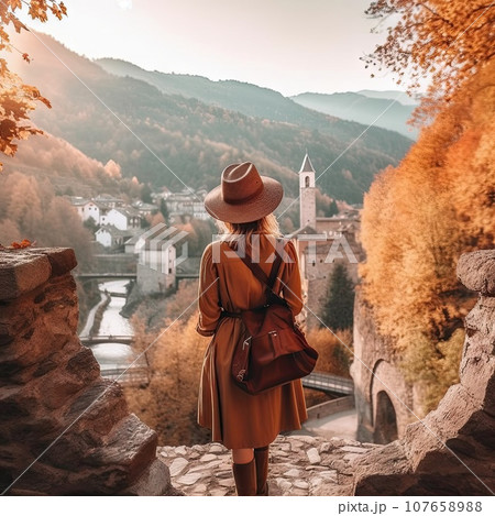 a woman in an orange coat and hat, standing on the edge of a cliff looking out over a river 107658988