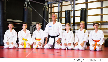 Portrait of boys and girls sitting on knees with male coach ready to taekwondo Portrait of boys and girls sitting on knees with male coach ready to taekwondo 107660189