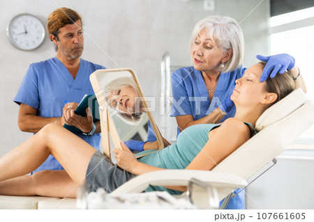Elderly cosmetologist with assistant planning facial procedure for female patient Elderly cosmetologist with assistant planning facial procedure for female patient 107661605