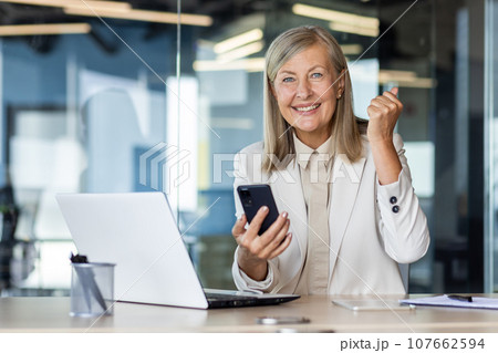 Portrait of a senior gray-haired businesswoman working in the office at a laptop, holding a phone in her hands, happy and smiling at the camera, showing a victory gesture with her hand. Portrait of a senior gray-haired businesswoman working in the office at a laptop, holding a phone in her hands, happy and smiling at the camera, showing a victory gesture with her hand. 107662594