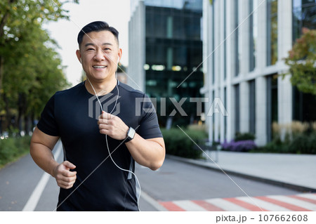 Portrait of an Asian young man running in headphones on a city street in the morning, smiling at the camera. 107662608