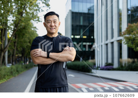 Portrait of a young Asian male sportsman standing on the street of the city in black sports clothes, hands folded on his chest and smilingly looking at the camera. 107662609