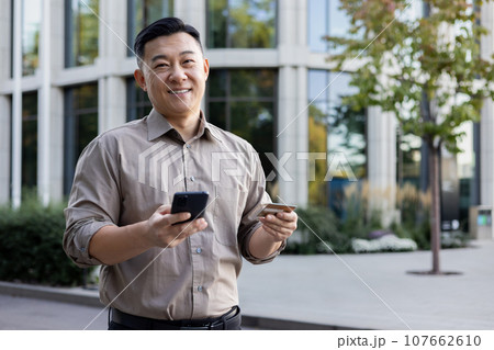 Portrait of a young Asian man standing on the street, holding a phone and a credit card, smiling and looking at the camera. 107662610