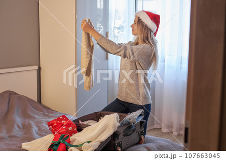 Christmas time. Woman puts knitted winter clothes and gifts in suitcase in room. Travel, vacation 107663045