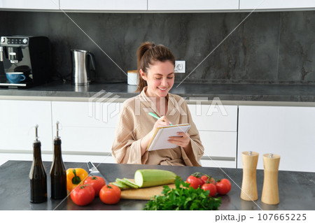 Portrait of beautiful, smiling young woman making list of meals, writing down recipe, sitting in the kitchen with vegetables, doing house errands 107665225