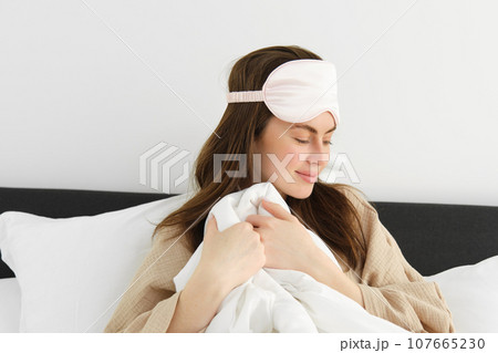 Tender and beautiful woman with sleeping mask, wakes up in morning, covers herself with duvet, smiles with gentle gaze aside, resting at home 107665230