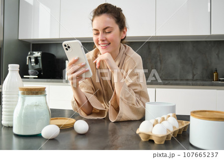 Attractive young cheerful girl baking at the kitchen, making dough, holding recipe book, having ideas 107665237