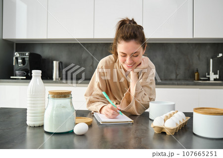 Attractive young cheerful girl baking at the kitchen, making dough, holding recipe book, having ideas 107665251