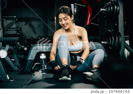 Woman tying the shoelace of her running shoe, holding a shaker bottle of water. The shot emphasizes the importance of staying hydrated during exercise. 107666148