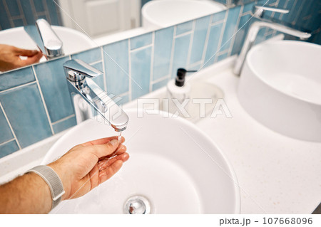 A man's hand touches the water in a beautiful sink with a metal faucet next to an mirror. Close-up of an elegant faucet in the bathroom sink next to stylish decorations.. High quality photo A man's hand touches the water in a beautiful sink with a metal faucet next to an mirror. Close-up of an elegant faucet in the bathroom sink next to stylish decorations.. High quality photo 107668096