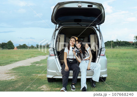 Two women friends and open trunk of car to sit back and relax after reaching rest place Two women friends and open trunk of car to sit back and relax after reaching rest place 107668240