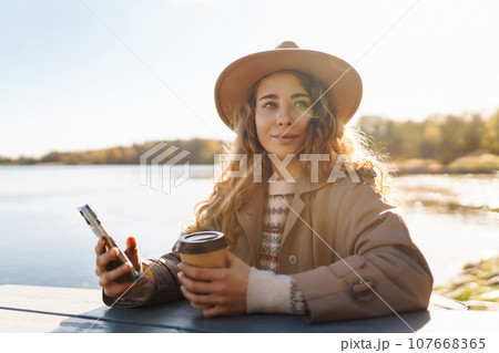 Smiling woman in hat on terrace with phone enjoys sunny autumn weather. Concept of relaxation, rest. Smiling woman in hat on terrace with phone enjoys sunny autumn weather. Concept of relaxation, rest. 107668365