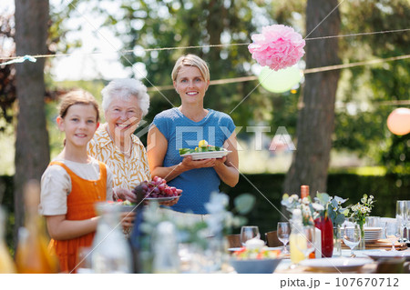 Grandmother, mother and daughter setting table for summer garden party. Three generations of women in family, bringing food and drinks at table. Grandmother, mother and daughter setting table for summer garden party. Three generations of women in family, bringing food and drinks at table. 107670712
