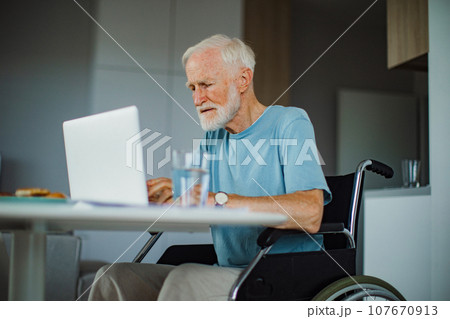 Senior man in wheelchair working from home during retirement. Elderly man using digital technologies, working on laptop. Concept of seniors and digital skills. Senior man in wheelchair working from home during retirement. Elderly man using digital technologies, working on laptop. Concept of seniors and digital skills. 107670913