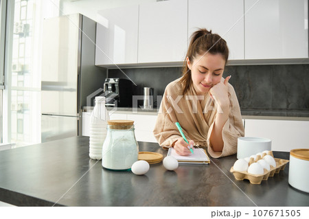 Attractive young cheerful girl baking at the kitchen, making dough, holding recipe book, having ideas Attractive young cheerful girl baking at the kitchen, making dough, holding recipe book, having ideas 107671505