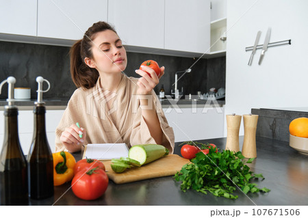 Portrait of beautiful smiling woman, writing her healthy menu, eating tomato while cooking, making grocery list, sitting in the kitchen Portrait of beautiful smiling woman, writing her healthy menu, eating tomato while cooking, making grocery list, sitting in the kitchen 107671506