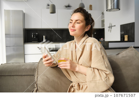 Beautiful young woman resting on sofa, enjoying her morning at home, holding glass of orange juice and smartphone, looking outside window 107671507