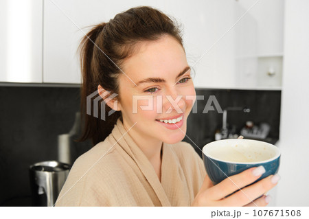 Portrait of good-looking young woman starting her day with cup of coffee, standing in the kitchen and drinking cappuccino from big mug, enjoying favourite drink in the morning 107671508